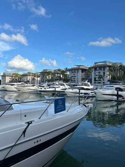 An outdoor, medium-shot, eye-level, full-color photograph depicts a marina with several luxury yachts docked in the water. The scene is located in the city of Ko Kaeo, Thailand. In the foreground, the bow of a white speedboat is prominently featured, with a blue sign attached to its railing that reads "BOAT IN THE BAY CHARTER & MANAGEMENT +66 800 40 40 59". The water is calm, reflecting the blue sky, white clouds, and the surrounding buildings and yachts.

In the mid-ground, a row of sleek, modern yachts are moored side-by-side. These yachts are predominantly white with dark accents. Behind the yachts, a line of multi-story buildings with beige facades, dark roofs, and balconies can be seen. Palm trees are interspersed among the buildings, suggesting a tropical or subtropical climate. The sky is a vibrant blue, dotted with fluffy white clouds. The lighting suggests it is daytime, likely morning or afternoon, with the sun casting soft shadows. There are no people visible in the frame.