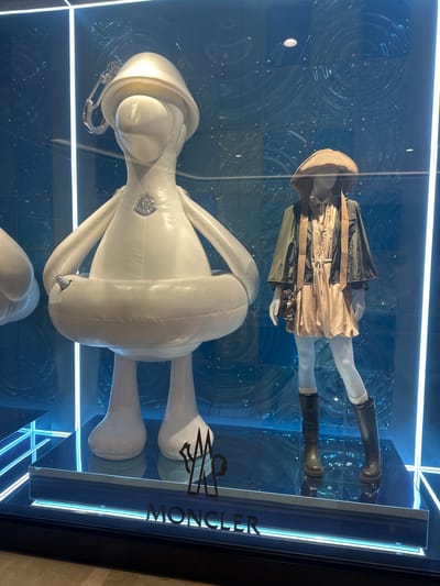 A shop window display features two mannequins and a large inflatable figure. The upper mannequin is dressed in a beige dress, a dark green jacket, a brown hat, and black boots with white socks. The mannequin is wearing white tights and holding a brown clutch. The lower figure is a large, white, inflatable character with round features, resembling a plush toy. It has a large round head with a hood, a thick torso, and long, rounded limbs. Both the mannequin and the inflatable figure have Moncler logos visible on them. The background of the display is a blue, textured surface with circular patterns and is illuminated by bright white lights. The Moncler logo is also prominently displayed on the right side of the shop window. The overall setting appears to be a retail store in Sydney, Australia. The time of day is not discernible, and the weather is not relevant to the indoor display.