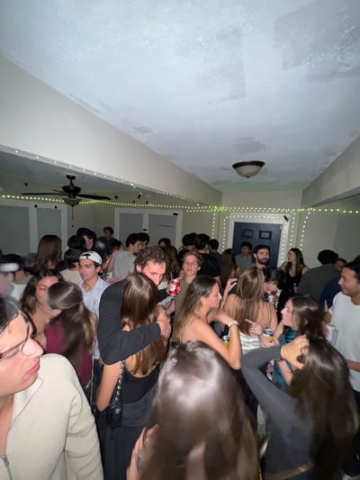 The image displays a crowded indoor social gathering in South Bend, United States, populated by approximately 30-40 young adults, primarily in their late teens to early twenties. The room features light-colored walls and a textured white ceiling. A dark-colored ceiling fan is visible on the left side of the ceiling, and a bowl-shaped light fixture is mounted centrally towards the right. Greenish-yellow LED string lights are strung along the perimeter of the room near the ceiling. Multiple door frames or openings are present in the background, including a distinct dark blue door. Individuals are standing in close proximity, indicating a high-density environment. Several participants are holding red and white beverage cans. Attire is casual, encompassing various shirts and tops. Observed interactions include standing, conversing, and some individuals embracing or with arms around others. The scene is illuminated by dim ambient light.