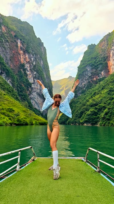 A woman stands on the bow of a boat on a body of water in Đồng Văn Commune, Vietnam, with arms raised and spread. The boat is white with a green astroturf surface. She is wearing a light green one-piece swimsuit, a light blue blouse, white knee-high socks, and gray sneakers. Her hair is pulled back. Behind her are tall, rocky mountains with green vegetation. The sky is partly cloudy.