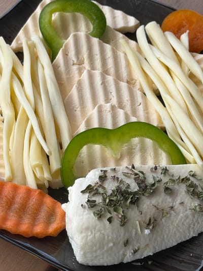 A close-up, overhead shot displays a platter of cheeses and vegetables. In the foreground, a block of white cheese, sprinkled with dried herbs, sits on the right. To its left, a wavy-cut slice of bright orange carrot is visible. In the middle ground, a ring of green bell pepper is positioned over a series of thinly sliced, pale yellow cheeses with ridged surfaces. To the left of these slices, several strands of string cheese lie in a bundle. In the background, a bright orange dried apricot is visible on the upper right, along with more of the ridged cheese slices and another green pepper ring. The food is arranged on a dark-colored, possibly ceramic platter, which is set on a wooden surface. The lighting suggests an indoor setting. The image captures a still life arrangement of food items, without any people or actions depicted. The image was taken in Byurakan, Armenia.