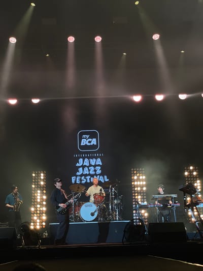 The image shows a musical performance on a stage at the "myBCA International Java Jazz Festival". The location is likely Salembaran, Indonesia, as the festival takes place there. Several musicians are visible: a drummer, a guitarist, a saxophone player, and a keyboardist. Stage lighting illuminates the performers and the festival backdrop.