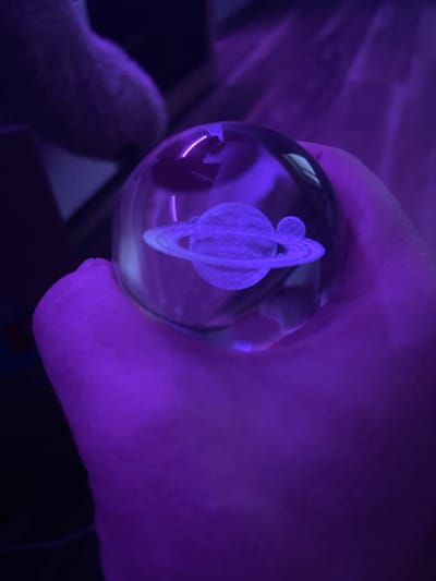 A hand, illuminated by purple light, holds a glass orb containing a miniature model of Saturn with a moon. The background is dark and blurred, with hints of purple light reflecting on surfaces. The image appears to be taken indoors, with no identifiable landmarks of Gliwice, Poland. The focus is on the orb and the hand.