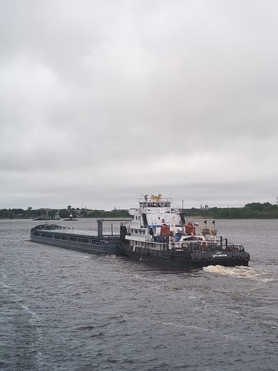 A medium shot depicts a tugboat pushing a barge on a river under an overcast sky. The tugboat is black and white, with multiple decks and a visible Russian flag. The barge is long and gray. The river surface is choppy, with visible wake behind the tugboat. In the background, the cityscape of Shestovo, Russia, is visible with vegetation and buildings.
