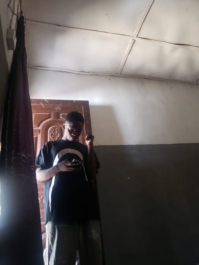 The image shows a person standing inside a room. The room has light-colored walls on the left and a darker, possibly gray wall on the right. A door with a patterned design is visible behind the person. The person is wearing a black shirt with a white stripe and green pants with the word "BELANCINGA" repeated on them. The lower part of the image is dark and obscures the details of the floor. There is insufficient information to determine the location of this scene in Atlanta, United States of America.