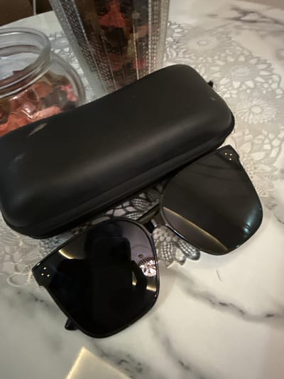 A black, rectangular sunglasses case rests on a white, marbled surface adorned with a delicate white lace doily. To the right of the case, a pair of black sunglasses is partially open. The sunglasses feature large, dark lenses and a thick black frame, accented with small gold embellishments on the upper corners of the lenses. A decorative element, possibly a small gem or crystal, is visible on the bridge of the sunglasses. In the background, a glass jar filled with dried red petals and another container with more dried floral elements are slightly out of focus, suggesting a decorative arrangement. The lighting is soft, creating subtle reflections on the sunglasses and the marble surface. This scene is set in an indoor environment, likely a table or countertop. The provided information states the location is the City of Pematang Siantar, Indonesia.