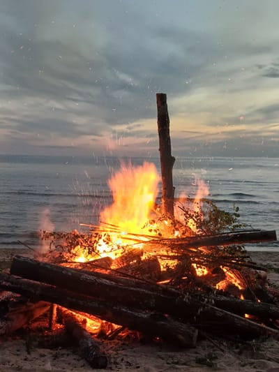 The media file depicts a bonfire on a beach at dusk. The scene is dominated by a large pile of logs engulfed in bright orange flames, casting a warm glow on the surroundings. Sparks and embers are seen rising from the fire, some of which drift towards the sky, creating a mesmerizing visual effect.

The background features a body of water, likely the Baltic Sea, with soft, pastel-colored clouds reflecting the fading daylight. The sky is a blend of gray, pink, and orange hues, indicating the transition from day to night. The beach is composed of sand and scattered driftwood. Small branches and leaves are visible within the burning logs, adding texture to the fire.

The image appears to be taken in Vārzas, Latvia. The overall mood is peaceful and serene, with a touch of wildness due to the intensity of the flames. The time of day is clearly evening, and the weather seems calm. There is no visible text in the image.