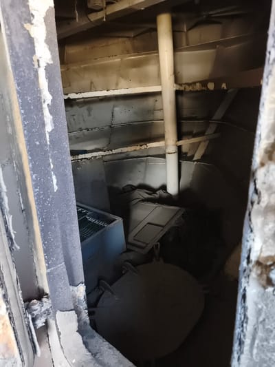The image depicts the interior of a structure, possibly a ship's hold or a storage area, with a strong sense of disuse and decay. The scene is dimly lit, with dust and debris coating most surfaces. Prominent objects include a large, rounded, dark-colored item in the foreground that appears to be a heavy container or tank, partially covered in dust. To its left, a light blue metal container with a grid-like opening sits on the floor. A white pipe runs vertically along the right side of the image, disappearing into the upper structure.
The walls and ceiling are constructed of metal panels, many showing signs of wear and damage, with peeling paint and rust. Structural beams are visible, adding to the utilitarian aesthetic. The overall impression is one of an abandoned or disused space, possibly a cargo hold or engineering section of a vessel given the industrial materials and confined nature. The limited visibility and the accumulation of dust suggest a lack of recent activity. No people are visible in the frame. The context of Chimbote, Peru, does not provide specific visual cues within the image itself, but it frames the location as a coastal industrial or port city. The image is a still photograph, and therefore, lacks information about time of day or weather conditions. There is no visible text in the image.