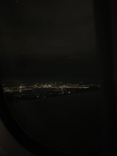 The image captures a dark, nocturnal landscape, likely viewed through a window, as suggested by the dark, curved frame visible in the bottom right. The primary subject is an elongated, vertical band of light that runs along the right side of the frame. This bright streak is composed of numerous small, warm-toned points of light, appearing to be streetlights or distant settlements.

A notable detail is the textured, somewhat jagged appearance of the main light band, which suggests that the light is being refracted or diffused through a surface, possibly a rain-streaked, smudged, or scratched window. This effect creates a "veiny" or "streaked" quality to the illumination. The surrounding areas are predominantly dark, implying a vast unlit expanse, typical of a rural or mountainous region at night.

Given the location context of Khánh Sơn, Vietnam, a mountainous district, this scene likely depicts lights from a winding road or scattered communities nestled within a valley or along mountain slopes, viewed in darkness. No specific activity, people, or visible text are present. The time of day is clearly night.