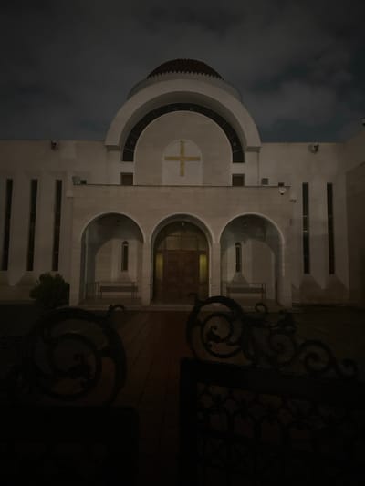 The image displays a grand, illuminated building at night, strongly resembling a church or religious institution, set against a dark, cloudy sky. The architecture is prominent, featuring a light-colored facade, likely stone.

On the left side of the structure, there is a distinctive semi-circular or domed section with several concentric tiers. A golden cross is centrally placed on a circular feature within this section, highlighting its religious significance. The main body of the building presents a series of symmetrical arched recesses or entranceways, through which warm artificial light emanates, suggesting interior illumination. Horizontal rectangular elements or windows are also visible across the facade.

To the right, a dark, ornate wrought-iron gate or fence partially obscures the view, characterized by intricate scrollwork and decorative patterns. A small amount of dark foliage is visible near the base of the building on the lower right.

The time of day is night, with the building brightly lit, creating a stark contrast with the dark, overcast sky. No human activity or visible text is present in the scene. The overall impression is one of solemnity and architectural grandeur in a nocturnal setting, consistent with the religious structures found in Egkomi Lefkosias, Cyprus.