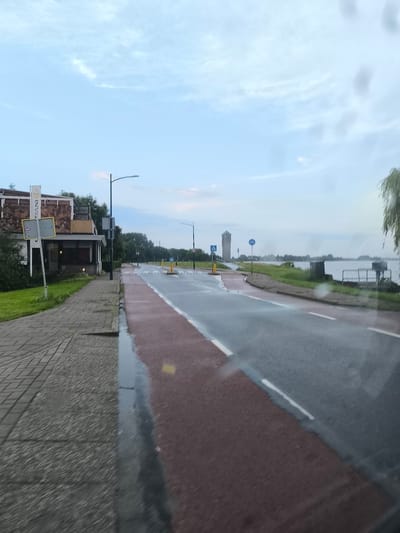 The image shows a street scene in Aalsmeer, Netherlands. A paved sidewalk runs along the left side of the road, with a building situated next to it. The building has a brown, textured facade and a white sign with text. Trees and greenery line the edge of the sidewalk. The road itself is asphalt with red and white markings, indicating lanes and a bike path. The road appears wet, suggesting recent rain. In the distance, a tall water tower is visible, and a body of water stretches out to the right, with a railing and some structures along its edge. The sky is overcast with a few scattered clouds. The scene depicts a typical street in a Dutch town, possibly near a waterfront.