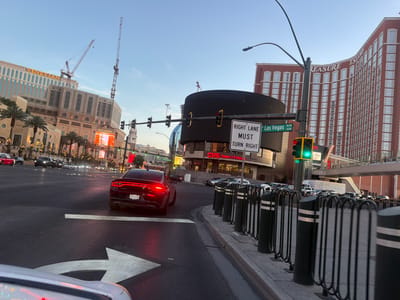 The image captures an urban scene in Paradise, Nevada, United States. The time of day appears to be early evening, as indicated by the sky's color. A black sedan, with license plate "NZ-230-878" is in the foreground, positioned on a street marked with a right-turn arrow. The car's brake lights are illuminated.
Visible structures include the Venetian and Treasure Island hotels, both iconic landmarks in Paradise. Construction cranes are near the Venetian, suggesting ongoing construction or maintenance. Street signs indicate "Las Vegas Blvd" and the street number "3249". A large electronic sign, partially obscuring the view, is present, along with a "Right Lane Must Turn Right" sign. A CVS store is also visible with the text "CVS Open 24 Hours."
Traffic lights are present, showing green on the right side and red on the other side of the intersection. A line of bollards is present near the sidewalk. Cars are parked along the street in the middle ground. The overall environment suggests a bustling urban intersection in a tourist area.