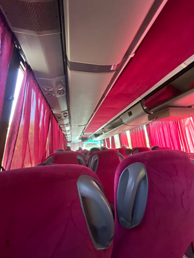 The image shows the interior of a bus, viewed from a low angle looking forward. The seats are upholstered in a bright pink fabric. Red curtains are drawn along the windows on the left side of the bus, with sunlight filtering through them. The ceiling of the bus is visible, with air vents and lighting fixtures. In the distance, the aisle of the bus is visible, with other passengers seated. The overall impression is one of a brightly lit and somewhat cramped interior, suggesting a journey in progress. The bus is located in Buhovtsi, Bulgaria.