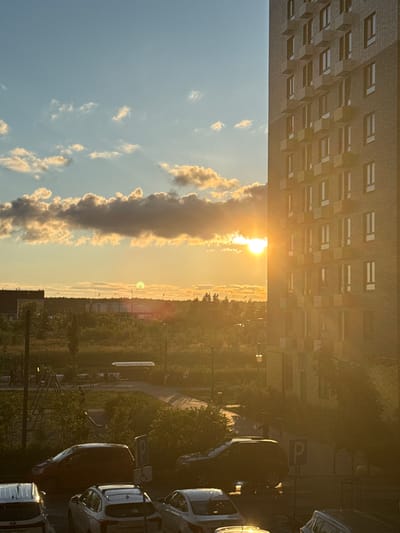The image depicts a sunset view from an apartment building in Drozhzhino, Russia. The sun is setting behind a treeline and some buildings in the distance, casting a warm glow over the scene. A tall, modern apartment building with visible windows and balconies occupies the right side of the frame. In the foreground, several parked cars are visible, along with a children's playground with a slide and climbing structure. The sky is a mixture of blue with scattered, golden-tinged clouds, illuminated by the setting sun. The overall environment appears to be a residential area with interspersed green spaces.