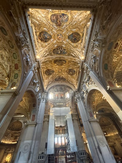 The image shows the interior of a grand church, likely a cathedral, in Bergamo, Italy. The perspective is from the ground looking upwards at the highly ornate ceiling and dome. The ceiling is a complex structure of golden ornamentation, frescoes depicting biblical scenes, and intricate architectural details. Large, fluted columns line the nave, supporting the arched ceiling and a central dome that lets in natural light. The overall impression is one of opulence and religious grandeur. The church appears to be well-maintained and possibly undergoing some minor restoration work, as indicated by the presence of some scaffolding or sheeting near the altar area. There are no people clearly visible in the image.