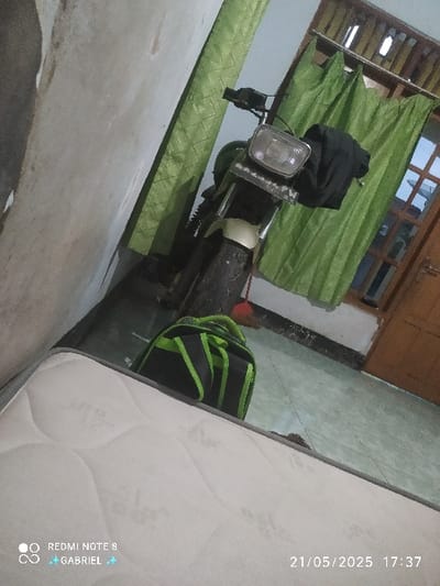 The image shows a green motorcycle parked in a room, partially obscured by a green curtain. A black backpack is on the floor near the motorcycle. There is a white mattress in the foreground of the image.  The location of the image is Kebumen, Indonesia, based on the coordinates -7.685 latitude and 109.681 longitude.  The image appears to be taken from a low angle and captures a portion of the room's interior. The camera phone watermark indicates the image was taken on 21/05/2025 at 17:37.  The image shows no people.