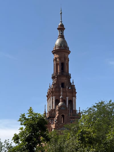 This is a close-up, low-angle shot of the Plaza de España in Seville, Spain, focusing on one of its prominent brick towers. The tower is elaborately decorated with tilework and has multiple levels with arched windows and decorative columns. It is topped with a conical spire. Lush green trees partially obscure the lower and mid-sections of the tower, contrasting with the bright blue sky above. The image captures a sunny day with clear visibility. No people or animals are visible in the frame.
