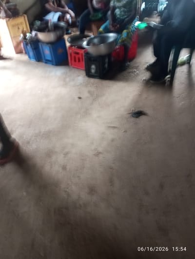 The image depicts an outdoor scene, likely in a marketplace or communal gathering space in Dokan Tofa, Nigeria. In the foreground, a person's legs and feet are visible, one wearing a red shoe. The ground is dirt. In the mid-ground, several individuals are gathered around tables and crates. To the left, a cardboard box with "Guinness" printed on it is visible. Blue and red plastic crates are stacked, some containing metal bowls. One person appears to be holding a green bottle. The scene is somewhat blurry, indicating motion or low light conditions. In the background, more people are seated and standing. The overall atmosphere suggests a casual, community-oriented setting.