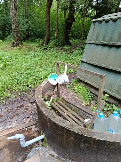 In the city of Staryi Pokrov, Russia, a natural spring emerges from the ground, channeled through a concrete structure. The spring is covered by a green, corrugated roof. Two large plastic water bottles with blue handles are positioned beside the concrete rim. A wooden railing, suspended by wooden posts, holds two containers: a white mug with blue markings and a green and white bucket. Adjacent to the spring, a PVC pipe with various connectors directs water into a puddle on the ground. The scene is set within a lush, green forest, with trees and foliage surrounding the spring. The ground around the spring is damp and muddy, with some wooden planks laid out. A birdhouse is visible attached to a tree in the background.