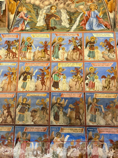 This image shows a section of frescoes in the Rila Monastery, Rilski manastir, Bulgaria. The frescoes are arranged in rectangular panels, depicting religious scenes with angels, demons, and human figures. Many panels feature text in what appears to be a Cyrillic alphabet. The style is Byzantine, characterized by flat figures, strong outlines, and symbolic colors. Some sections show damage and deterioration, particularly towards the right side of the image, revealing signs of age and wear.