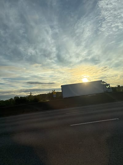 The image captures a side view of a white, refrigerated truck driving on a highway. The truck is positioned on the right side of the frame, moving from left to right. The sun is low in the sky, creating a bright glare that obscures parts of the truck and the horizon. Wispy clouds are visible in the sky, some of which are illuminated by the sun. The highway itself is dark asphalt with white lane markings. The surrounding environment appears to be rural, with a line of trees and foliage visible beyond the highway embankment. There are also some yellow and blue signs on the embankment, one of which appears to be a warning sign. The overall lighting suggests either sunrise or sunset. The scene is likely taking place on a major road or motorway in South Staffordshire, United Kingdom, given the context provided.