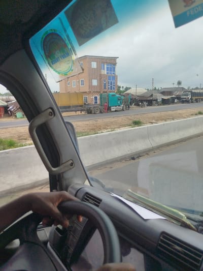 The image captures a view from inside a vehicle, looking through the windshield. The driver's hand is visible on the steering wheel. The dashboard has a piece of paper and what appears to be a small green item on it. Outside the vehicle, a road is visible with several trucks and buildings lining the sides. One building is a multi-story structure with blue-tinted windows. There are also smaller structures with corrugated metal roofs. The sky is overcast. The scene is consistent with a roadside view in Odiderengwaji, Nigeria, given the presence of typical structures and vehicles found in such locations.