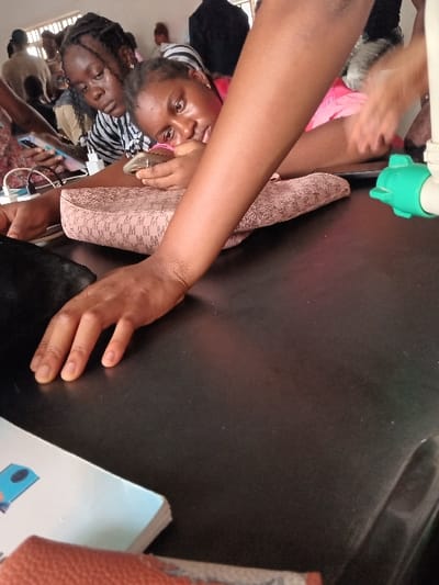 The scene is set indoors in OGBOKO, Nigeria, at what appears to be a communal or educational facility, observed from a low vantage point across a dark, smooth tabletop. In the midground, two young women are positioned. The woman closer to the right, wearing a pink top, rests her left forearm on a beige patterned purse or bag. She holds a smartphone horizontally in her right hand, her attention directed towards the device or a point just beyond it. Behind her and to the left, another woman in a black and white striped top holds a smartphone in her left hand, also looking downwards. Her hair is styled in dark braids. Multiple white charging cables and power adapters are visible on the table near these individuals, indicating device charging. A light-skinned forearm and open hand extend from the right foreground, resting on the tabletop. On the far right, a white pipe with a green connector is visible just above the table surface. In the bottom-left corner, a light blue and white book is partially visible, resting on an orange-brown textured object. The background features several out-of-focus individuals and a window with vertical bars, suggesting an interior space with ambient lighting.