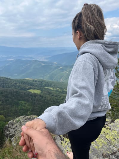 The image captures a close-up of two people holding hands while standing on a rocky outcrop, overlooking a vast mountainous landscape. The orientation of the image is vertical.
The subjects are two individuals, one seen from the back, wearing a gray hoodie with white text on the sleeve that appears to read "Catch Wav 1873". This person has light brown hair tied back and is wearing black leggings. Their hand is being held by another person, whose hand is visible, displaying dark hair and a fingernail with red polish. The focus is on their clasped hands, suggesting a connection or shared experience.
The setting is a dramatic natural environment. The background reveals a panorama of lush green mountains under a cloudy blue sky. The texture of the rocks in the foreground, with patches of green lichen, indicates a rugged, natural terrain. The scene evokes a sense of adventure, intimacy, and a connection with nature, possibly at a high altitude. The presence of both clear sky and clouds suggests variable weather conditions, but the overall lighting indicates it is daytime.
The image seems to capture a moment of shared trust and affection between the two individuals, perhaps a couple, enjoying a scenic view from a mountain peak. The visible text on the hoodie might be related to a brand or a personal message. The location cues of mountains and rocky outcrops are consistent with a high-altitude natural setting, and the mention of Simeonovets, Bulgaria, confirms a mountainous region.