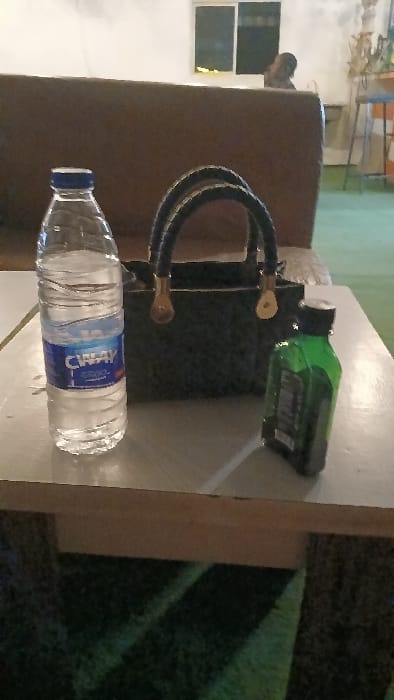 A water bottle with a blue cap and label sits on a white surface. Behind it is a hookah with black hoses and brass-colored connectors. A green bottle partially overlaps the hookah. These items are set on a table beside a brown upholstered chair. A window is visible on the left. The table surface is adjacent to a patch of green foliage or grass.
