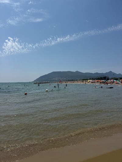 The image shows a crowded beach and a body of water on a clear, sunny day. In the foreground, the shallow water laps against a sandy shore. Further out, people are swimming and wading. The water is relatively calm with some small waves. In the mid-ground, a line of buoys and floats mark a swimming area. Beyond the water, a long stretch of beach is lined with numerous colorful umbrellas and beach chairs, indicating a popular tourist destination. People are visible lounging under the umbrellas and walking along the shore. In the background, a large, green, mountainous landscape rises above the beach. The sky is a clear blue with some scattered white, wispy clouds. The scene suggests a typical summer day at a busy coastal resort. Based on the mountainous backdrop and the popular beaches, this is likely the coastline near San Felice Circeo, Italy, a town known for its scenic beauty and its location on the Tyrrhenian Sea, nestled at the foot of Monte Circeo.