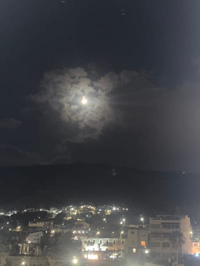 The image shows a night scene of Tiberias, Israel, with a full moon partially obscured by clouds. The moon is located in the upper center of the frame, casting a soft glow on the surrounding clouds. Below the moon, the city of Tiberias is visible, with numerous lights twinkling from buildings and streetlights. The city is built on a hillside, with structures cascading down towards the foreground. In the lower portion of the image, some buildings and palm trees are visible, suggesting an urban environment. The overall atmosphere is tranquil and serene, with the moonlight illuminating the night.