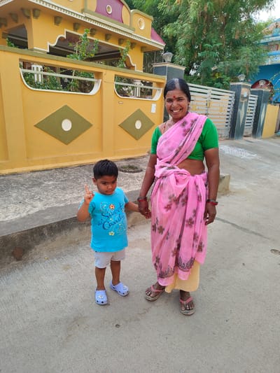 In Kammavaripalli, India, a child in a blue shirt and light-colored shorts stands next to an adult woman wearing a green blouse and a pink sari. The child holds up one finger. They are both holding hands. They are on a concrete road in front of a yellow house with a decorative wall. A grey gate and white designs on the road are visible in the background.