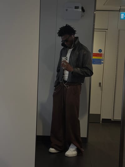 In a public restroom, likely within a transportation hub, a young Black man stands before a large mirror, taking a full-body selfie. He is dressed in a dark, possibly leather, bomber jacket worn over a white t-shirt, with dark brown, wide-leg sweatpants. White sneakers complete his outfit. He wears sunglasses and has earbuds in, while holding his smartphone in his right hand. Several rings adorn his fingers, and a bracelet is visible on his left wrist. The restroom's features include a white air freshener unit mounted on the wall, a white door with a blue and white directional arrow, and safety signs with pictograms. The floor is tiled in dark grey. The overall lighting is dim.