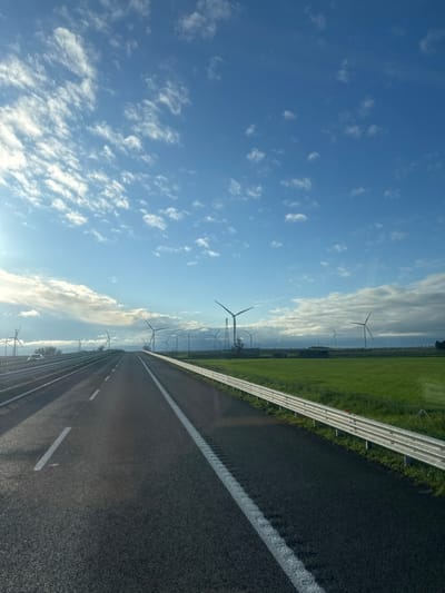 The image captures a multi-lane asphalt road, likely a highway, extending into the distance in Carapelle, Italy. The perspective is from a moving vehicle. On the left, at least two lanes are visible, separated by solid white lines, with a barrier bordering the far left and a light-colored car traveling in the same direction. On the right, a single lane is separated from a verdant agricultural field by a silver-colored metallic guardrail. A wind farm occupies the mid-ground and background, featuring numerous three-bladed wind turbines distributed across the landscape, with one prominent large turbine located slightly right of center. A tall lattice transmission tower stands near one of the larger turbines on the right. The sky is predominantly blue with scattered white cumulus clouds, and a bright light source, consistent with a low sun, is present on the far left, creating a glare. No individuals are visible in the scene.