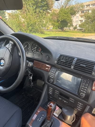 The image shows the interior of a parked car, looking out towards a residential street in Bozhurishte, Bulgaria. The car's dashboard is visible, with a steering wheel, instrument cluster, air vents, and a car stereo. A set of keys hangs from the ignition. Outside the car, trees and a multi-story apartment building can be seen. The scene is well-lit by daylight, suggesting it is daytime. The focus is on the car's interior, with the exterior serving as a backdrop.