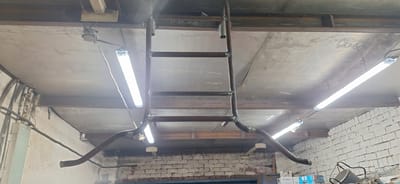 The image depicts an indoor scene, likely a workshop or industrial space, in Nakhodka, Russia. The focus is on a newly constructed metal structure resembling a ladder or scaffold, suspended from the ceiling. This structure appears to be made of welded metal pipes.

Two long fluorescent lights are mounted on the ceiling, illuminating the area. One light is on the left side of the frame, and another is on the right. The ceiling is a rough, grey concrete surface, supported by visible metal beams. The walls in the background are constructed of white brick. Electrical conduits and wires are visible running along the ceiling and walls, suggesting an active workspace.

The construction of the metal structure suggests an ongoing project or recent installation. The lighting is bright, indicating it could be daytime or the artificial lighting is sufficient for work. There is no visible human presence in the image.

A piece of text is faintly visible on one of the metal beams near the top of the frame: "51-212.001-12". This is likely a label or part number related to the construction or equipment in the space. The overall impression is one of a functional, utilitarian environment.