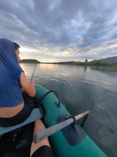 The image depicts a serene scene of two individuals fishing from an inflatable boat on a wide river. The person in the foreground, wearing a blue hooded shirt and black shorts, is actively fishing with a rod extended over the water. They are seated in the boat, which is predominantly green. Another individual, partially visible in the background, is also in the boat. The river flows calmly, reflecting the overcast sky. The background features a lush, tree-lined shore with rolling hills and mountains in the distance. The time of day appears to be late afternoon or early evening, given the soft, diffused light. The location is identified as the city of Ust-Buren, Russia, suggesting a natural, possibly rural, setting within this region.
