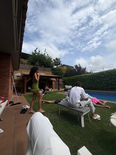 A group of people is gathered in an outdoor setting, likely a backyard in Sant Andreu de Llavaneres, Spain. There are individuals sitting on lawn chairs near a swimming pool, while others are standing and talking. A woman in a green dress is prominent in the foreground. The backyard includes grass, trees, a brick wall, and a building with a tiled roof. The sky is partly cloudy.