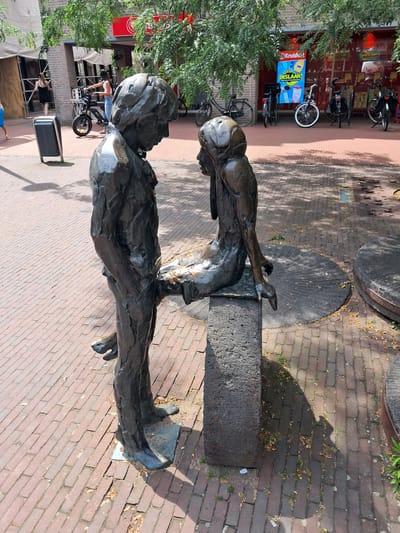 This image depicts a bronze sculpture of a young boy and girl in a seated position, positioned on a stone bench in the city of Veghel, Netherlands. The sculpture is situated on a brick-paved walkway in front of a building with a red facade, possibly a commercial establishment. The boy stands to the left of the girl, with his right leg extended forward and his left leg bent. The girl sits on the bench to his right, with her legs crossed and her right arm resting on her lap. Both figures appear to be in mid-conversation, with their heads turned towards each other. The environment suggests an outdoor public space, with trees providing shade and bicycles parked in the background. The scene is well-lit, indicating daylight.