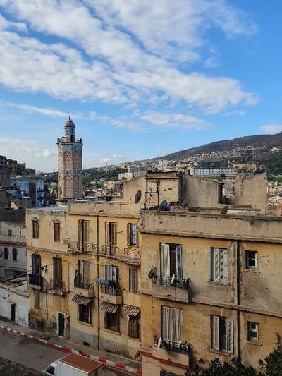 The image shows a tall, slender, ornate minaret of a mosque, rising up towards a clear blue sky with white puffy clouds. The minaret, with a green dome, is situated on a hillside in the city of Oran, Algeria, at coordinates 35.705 latitude and -0.65 longitude. It is flanked by a tall, aging yellow building with multiple windows, some with shutters, and several satellite dishes. The building, with peeling paint and visible bricks, appears to be several stories high, and has a balcony that appears to be lined with laundry hanging to dry. The image is taken from a lower angle, looking upwards towards the minaret. The background reveals a cityscape with buildings and houses spread across the hillside. The overall impression is one of an urban landscape with a mix of old and new architecture.