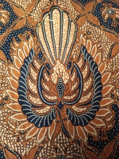 The image features an intricate batik fabric pattern. The central motif resembles a stylized bird or floral arrangement in shades of brown, tan, and navy blue. Small white dots accent the design, particularly around the edges of the navy blue sections. A gold metallic thread outlines many of the shapes, adding a subtle shimmer. The background is a mix of brown and navy blue with scattered tan and white dots. Overall, the pattern displays a traditional and ornate aesthetic.