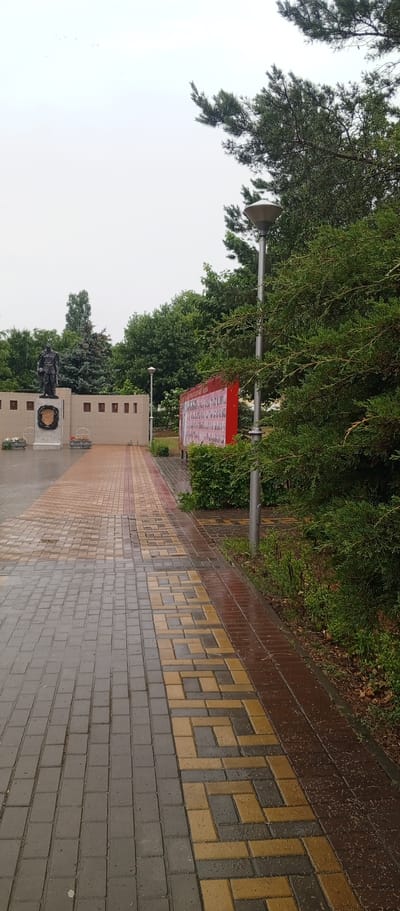 A memorial complex in Zhirnovsk, Russia, features a statue of a military figure on a pedestal, adorned with a wreath. Adjacent to the statue is a wall with several square indentations, likely for plaques. A patterned walkway, made of grey, red, and yellow interlocking pavers, leads towards the memorial. To the right of the walkway, a large red display board filled with numerous photographs is mounted on a pole. Dense green trees and bushes surround the area, and a lamppost stands near the display board. The ground appears wet, suggesting recent rain. The sky is overcast and pale.