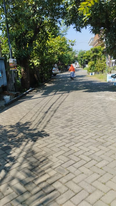 A person wearing an orange hijab and a blue skirt walks away from the camera down a paved residential street in Tanjungsari, Indonesia. The street is lined with green trees, and houses are visible on both sides. Sunlight casts shadows of the trees onto the paved road. A silver car is parked on the left side of the street.