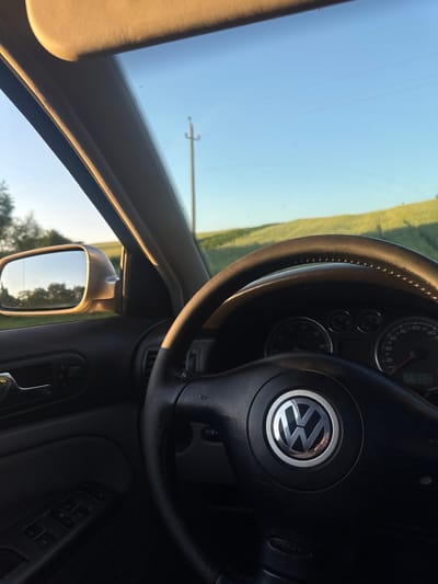 The image is an interior shot of a car, likely a Volkswagen, with the steering wheel and dashboard prominently displayed. The steering wheel has the distinctive Volkswagen logo in the center. To the left of the steering wheel, the car's window offers a view of a bright, clear blue sky and a green, rolling landscape, suggesting it's daytime and the weather is fair. A utility pole is visible in the distance. The interior of the car appears to be well-maintained, with the dashboard and door panel details visible. The side mirror reflects trees and the sky. The overall impression is of a relaxed drive through a rural setting.