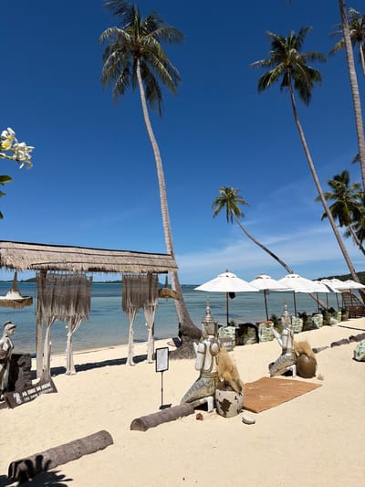 In Baan Thong Krut, Thailand, a serene beach scene unfolds under a clear blue sky. Palm trees, some leaning precariously, line the shore, their fronds swaying gently. The sand is light and grainy, leading to the tranquil turquoise water. A thatched-roof structure with macrame curtains provides shade, and nearby, white umbrellas are set up, suggesting a resort or dining area. Two ornate, seated statues, possibly of local deities, are positioned on the sand, adorned with vases and dry grass. Further down the beach, more umbrellas and seated beanbag-like structures are visible, creating an inviting atmosphere. A signpost indicates "Au Bout Du Monde - The Moon Experience," hinting at a specific establishment. The overall impression is one of peaceful relaxation in a tropical paradise.