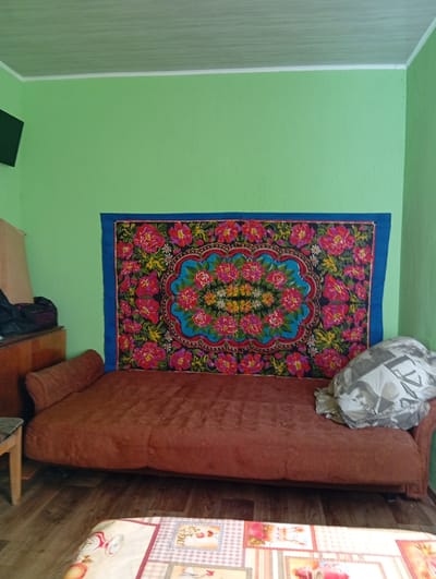 A couch with a red-brown fabric is positioned against a bright green wall. A large, colorful tapestry featuring a floral pattern is hung on the wall above the couch. A pillow with a black and white image of a person is placed on the right side of the couch. To the left of the couch, a wooden dresser is visible with some bags placed on top. A small wooden stool is to the left of the dresser. The floor is made of wooden planks. In the foreground, a table or surface covered with a patterned cloth is partially visible. The room appears to be a living space.