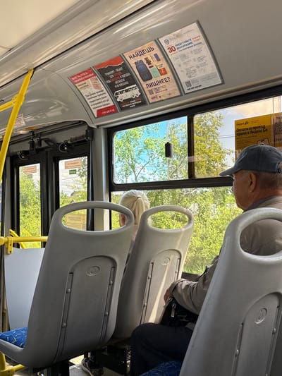 The image depicts the interior of a bus in Pankovka, Russia. Sunlight streams through the windows, illuminating the worn plastic seats and the advertisements affixed to the ceiling. A man wearing a baseball cap and a light-colored jacket is seated, facing away from the camera, looking out the window. A woman with blonde hair pulled into a ponytail is partially visible in the foreground, also seated. The bus appears to be in motion, with green trees and foliage visible outside the windows, suggesting a summer or spring day. Advertising posters are prominently displayed, including one for "Yandex Go" offering payment for travel via QR code. Other posters display information about insurance, taxes, and safety notices. The overall impression is a typical scene on public transportation.