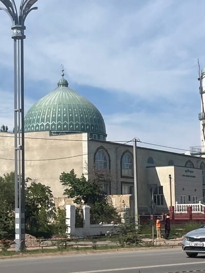 The image shows a street view in Balykchy, Kyrgyzstan, under a clear blue sky with some scattered clouds. The scene features a large, light green domed building that appears to be a mosque, with intricate decorative patterns along its dome. In the foreground, a dark metal structure, possibly part of a street light or signage, is visible. Adjacent to the domed building is another building with arched windows and light-colored walls. A road runs alongside the buildings, with a white car parked on the side. Further down the road, a person wearing an orange vest is visible, possibly working on or near the road. Trees and greenery are interspersed between the buildings and along the roadside. The overall impression is a daytime scene in a developed area.