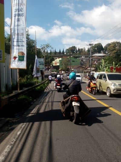 The image depicts a daytime street scene in Cipanas, Indonesia, under a clear blue sky with scattered white clouds. The perspective is from the rear of a motorcycle, looking forward down a multi-lane asphalt road.

In the immediate foreground, the rear of a dark-colored motorcycle is visible, carrying a rider wearing a black quilted jacket and a light blue full-face helmet. A dark, possibly canvas, bag is draped over the motorcycle's rear seat and fender. Shadows from overhead structures or poles are cast across the road surface.

To the left of the road, a metal guardrail runs parallel to the asphalt, behind which is dense green foliage. A tall, vertical white banner is affixed to a pole, displaying text that includes "CILUFIA CIFANAS AXI", "Lantai 2", "13.03.26", and pricing information "Rp 30k/35k/40k". The banner also features a graphic of a cartoon character with a cup of popcorn. Further along the left shoulder, a pedestrian in a white top walks away from the camera.

The road features a solid yellow line indicating a division between traffic lanes and a broken white line marking the left shoulder. A queue of multiple motorcycles, each with a rider wearing a helmet, is visible ahead in the left lane. In the right lane, a distinctly brightly colored (orange, red, yellow) custom motorcycle with a rider in a dark uniform is visible, alongside a light-colored minivan (possibly beige or champagne). A white car is also present further ahead in the left lane.

In the background, a hillside rises, featuring various buildings with facades in colors such as green and red, interspersed with numerous trees and utility poles with overhead wires.