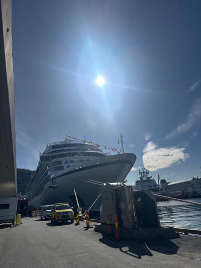 A large cruise ship is prominently docked at a concrete pier under a bright, clear blue sky. The image captures a sunny day, likely midday or early afternoon, with the sun creating a strong lens flare on the left side of the frame.
The majestic white and grey cruise ship features numerous decks lined with windows and balconies, showcasing its immense scale. Colorful signal flags are visible flying from a mast near its bow. On the concrete pier alongside the ship, several vehicles are parked, including a distinct yellow utility truck and a white service truck, possibly identified as part of "Maskin AS" from faint text on its side. Safety barriers with red and yellow stripes are positioned near the ship's hull, and some equipment marked "300C" and "JONZ" is also visible.
Across the calm, reflective water, other ships and buildings of a port city are discernible. Lush, tree-covered mountains rise in the distance, indicative of a fjord landscape, consistent with Bergen, Norway. The scene depicts a routine port call for the cruise ship. No identifiable people are clearly visible.